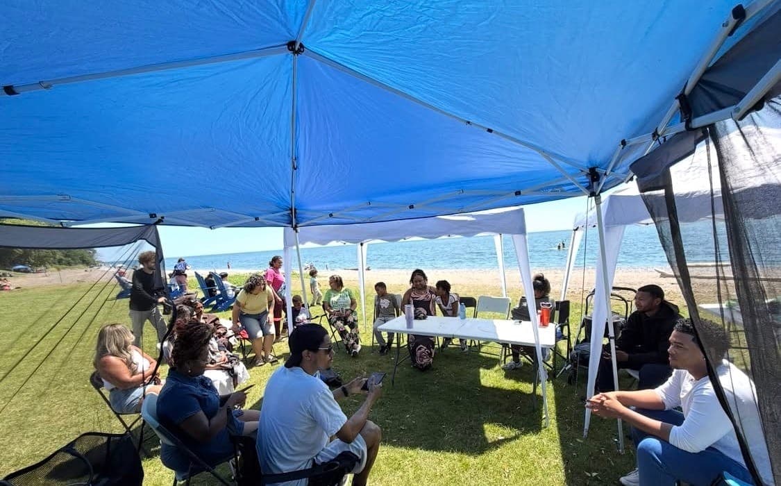 Members of Acts House sitting together in conversation under a canopy by the water.